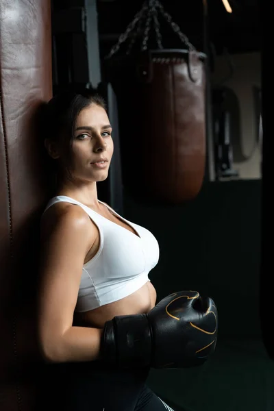 Boxeadora posando con saco de boxeo, sobre fondo oscuro. Concepto de mujer fuerte e independiente — Foto de Stock