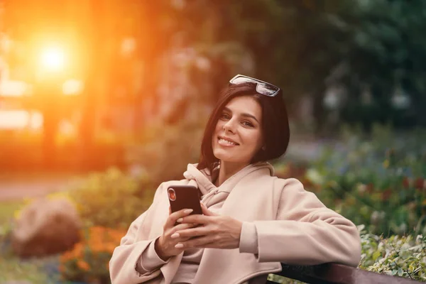 Menina feliz usando um telefone inteligente em um parque da cidade sentado em um banco — Fotografia de Stock