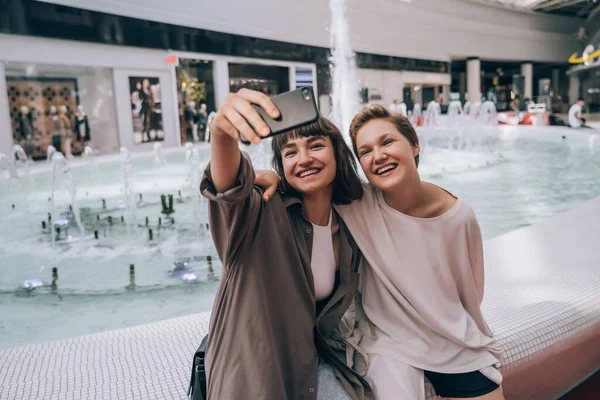 Dvě holky si berou selfie v obchoďáku, fontánu v pozadí — Stock fotografie