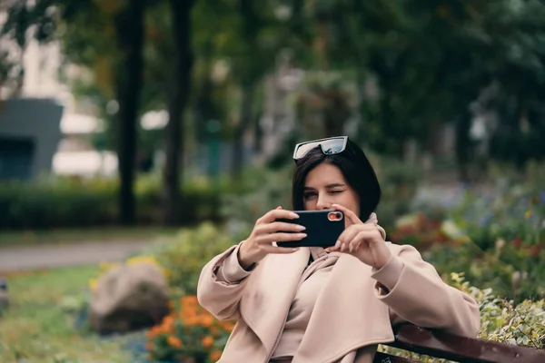 Menina feliz usando um telefone inteligente em um parque da cidade sentado em um banco — Fotografia de Stock