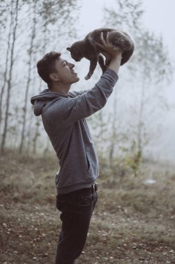 Adam parkta bir kedi tutuyor. Park sisle örtülü.