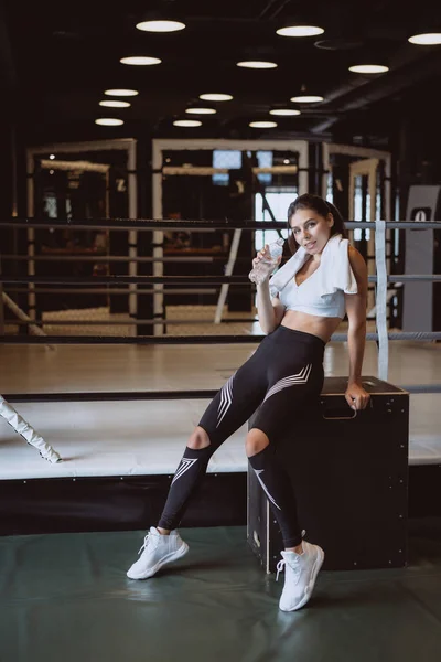 Hermosa joven con una toalla en los hombros bebiendo agua de una botella en el gimnasio — Foto de Stock