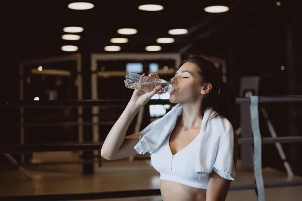 Linda jovem com uma toalha nos ombros bebendo água de uma garrafa no ginásio — Fotografia de Stock