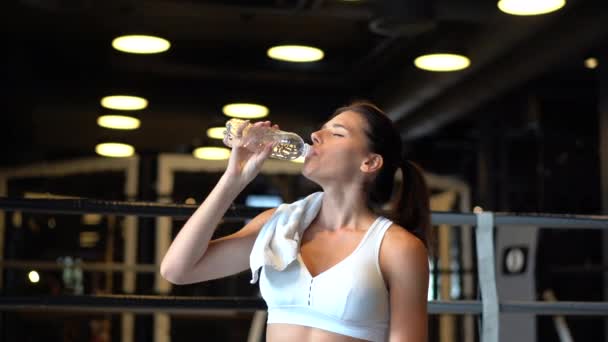 Прекрасная молодая женщина с полотенцем на плечах пьет воду из бутылки в спортзале — стоковое видео