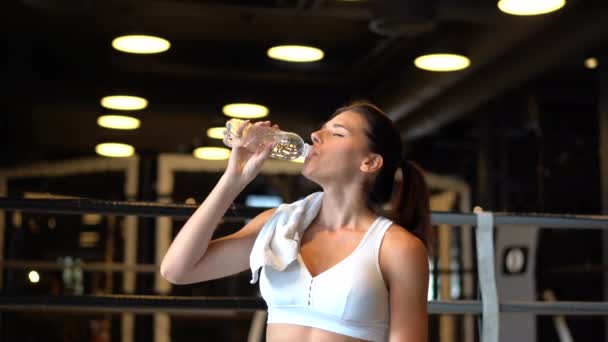 Hermosa joven con una toalla en los hombros bebiendo agua de una botella en el gimnasio — Vídeo de stock