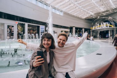İki kız alışveriş merkezinde selfie çekiyor, arka planda bir çeşme.