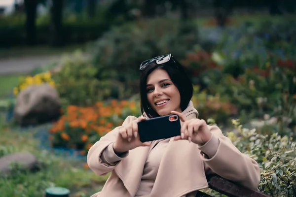 Happy girl za pomocą smartfona w parku miejskim siedzi na ławce — Zdjęcie stockowe