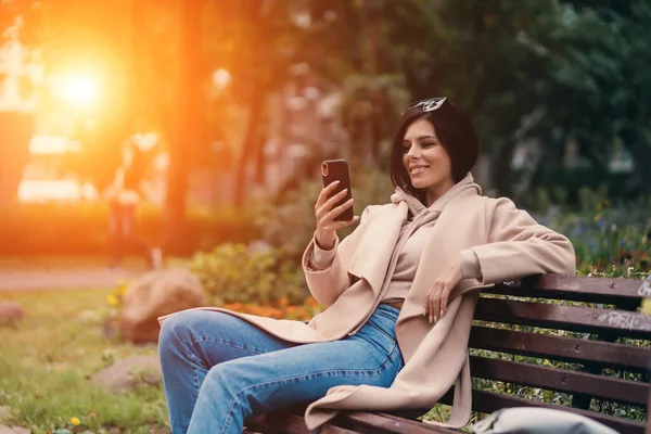 시립 공원에서 스마트폰을 사용하여 벤치에 앉아 있는 행복 한 소녀 — 스톡 사진