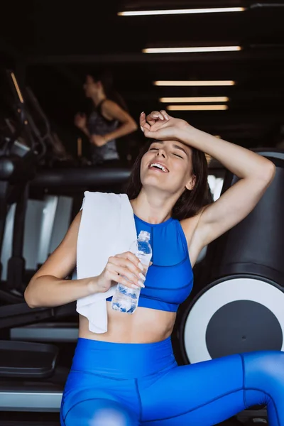 Hermosa joven con una toalla en el hombro bebiendo agua de una botella en el gimnasio — Foto de Stock