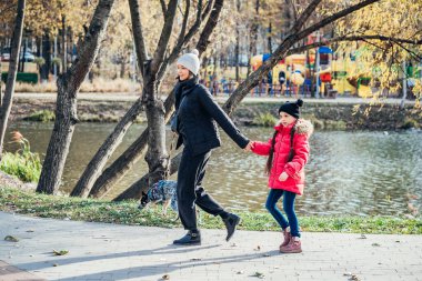 Mutlu anne ve kızı güz parkında köpekle yürüyor.