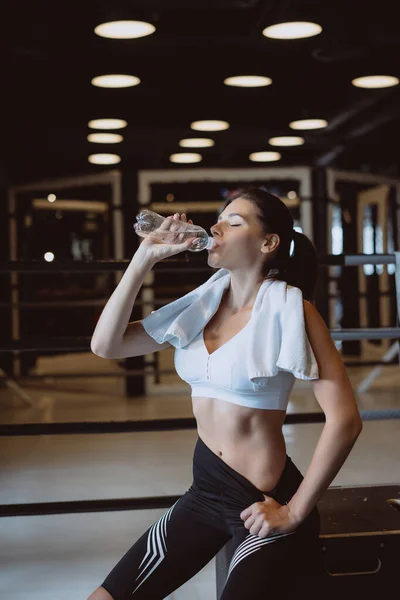 Wanita muda cantik dengan handuk di pundaknya minum air dari botol di gym — Stok Foto