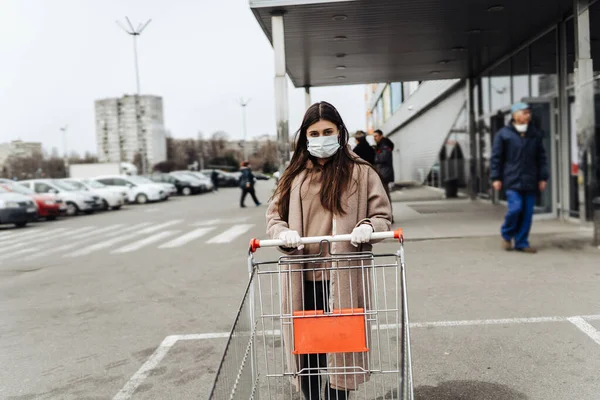 Молодая женщина в защитной маске против коронавируса 2019-nCoV толкает корзину. — стоковое фото