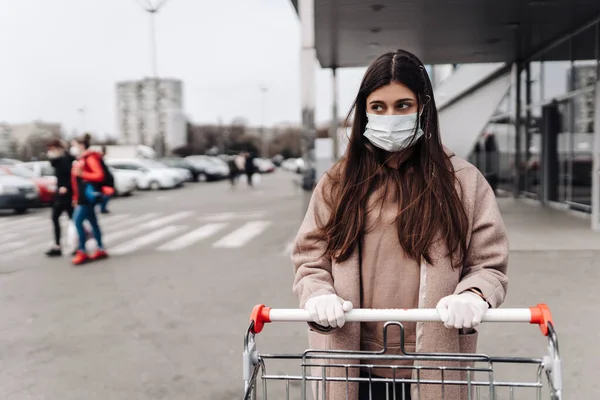 Молодая женщина в защитной маске против коронавируса 2019-nCoV толкает корзину. — стоковое фото