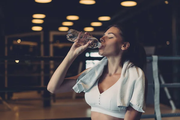 Linda jovem com uma toalha nos ombros bebendo água de uma garrafa no ginásio — Fotografia de Stock