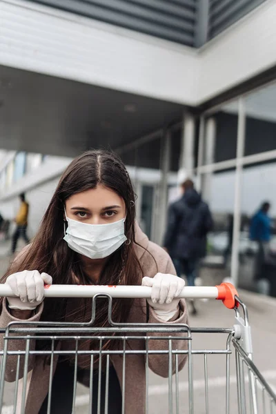 Mladá žena v ochranné masce proti koronaviru 2019-nCov tlačí nákupní košík. — Stock fotografie