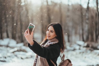 Mutlu gülümseyen genç kız ya da genç kadın kış parkında akıllı telefonla selfie çekiyor.