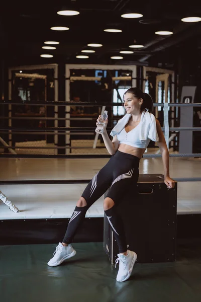 Prachtige jonge vrouw met een handdoek op haar schouders drinkwater drinken uit een fles in de sportschool — Stockfoto