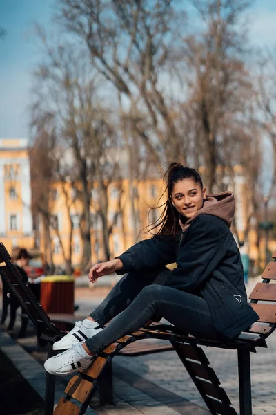 Fiatal nő ül padon a parkban, mosolyog és belenéz a kamerába — Stock Fotó