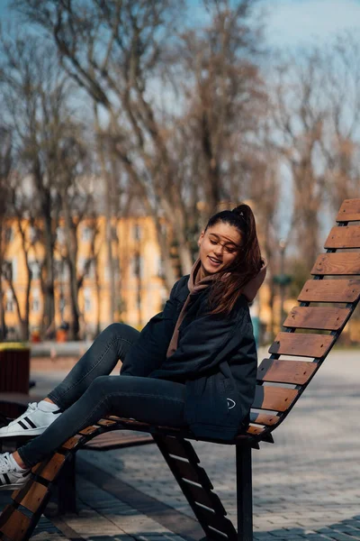 Mladá žena sedí na lavičce v parku, usmívá se a dívá se do kamery — Stock fotografie