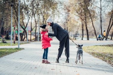 Mutlu anne ve kızı güz parkında köpekle yürüyor.
