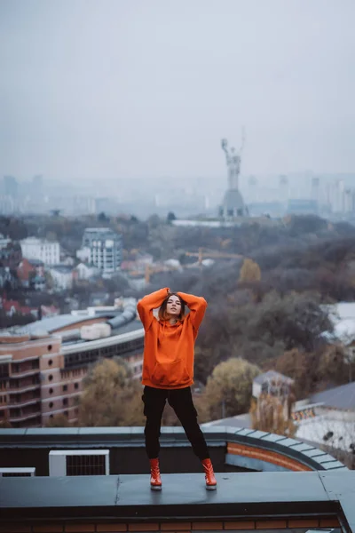 Menina em uma jaqueta laranja posa no telhado de um edifício no centro da cidade — Fotografia de Stock