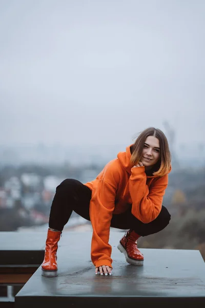 Chica con una chaqueta naranja posa en el techo de un edificio en el centro de la ciudad — Foto de Stock