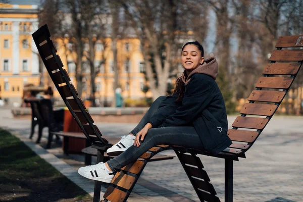 Mladá žena sedí na lavičce v parku, usmívá se a dívá se do kamery — Stock fotografie