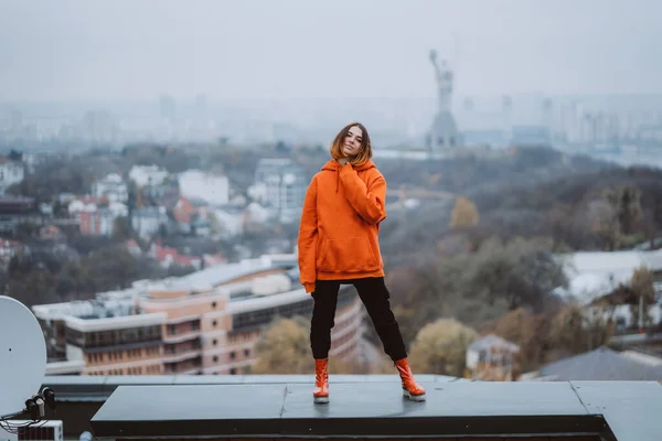 オレンジのジャケットを着た女の子が市内中心部の建物の屋根にポーズをとっています — ストック写真