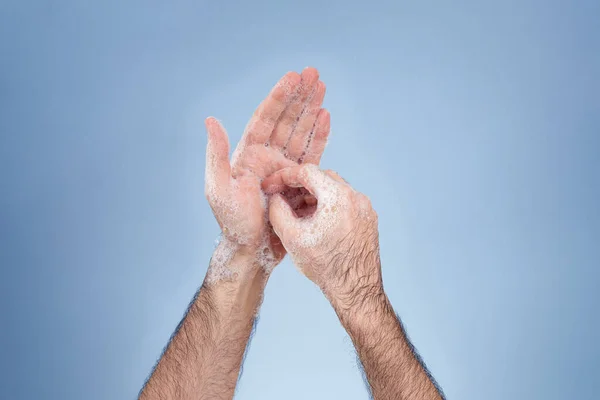 Hand washing with soap suds to achieve good prevention and hygiene. Mature man's hands with hairy arms. Close-up hands and smooth blue background.
