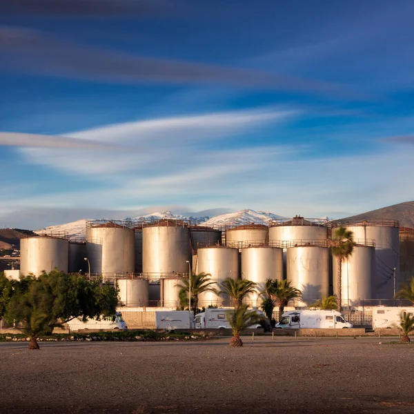 Metallic fuel tank storage plant. Sandy beach in the foreground, medium fuel tanks and snowy mountains in the background.