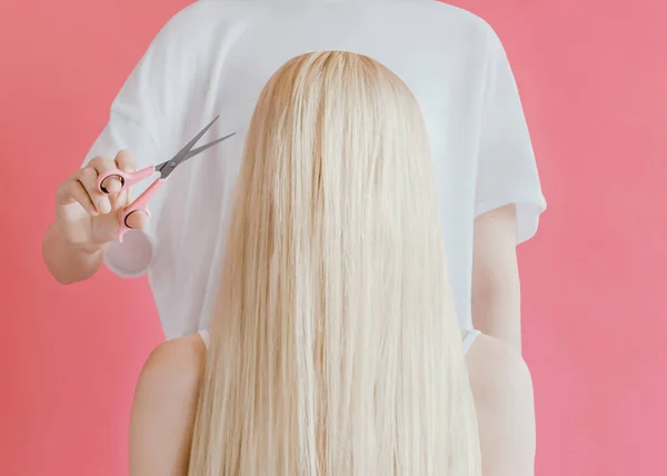 Hairdresser Design concept in minimal style Barber with scissors in her hands is standing behind a girl with long blond hair against pink background