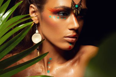 Bronz tenli, bronz tenli, makyajlı bir kadının yakından çekilmiş portresi. Yüzüne tropik orman boyası. Terli palmiye ağacı vahşi stili, egzotik aksesuar, seksi model..