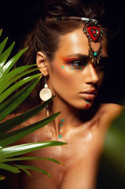 Bronz tenli, bronz tenli, makyajlı bir kadının yakından çekilmiş portresi. Yüzüne tropik orman boyası. Terli palmiye ağacı vahşi stili, egzotik aksesuar, seksi model..