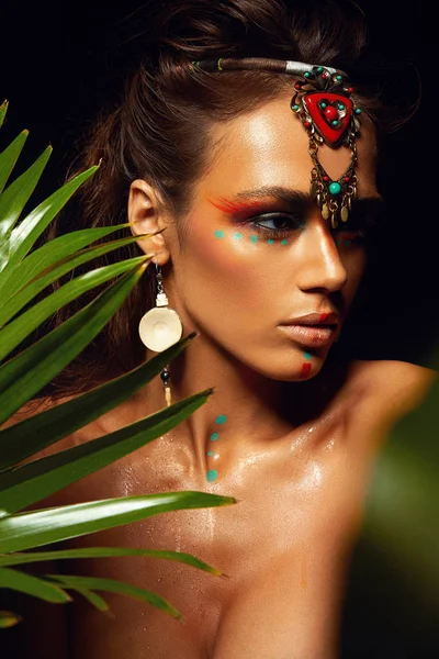 Bronz tenli, bronz tenli, makyajlı bir kadının yakından çekilmiş portresi. Yüzüne tropik orman boyası. Terli palmiye ağacı vahşi stili, egzotik aksesuar, seksi model..