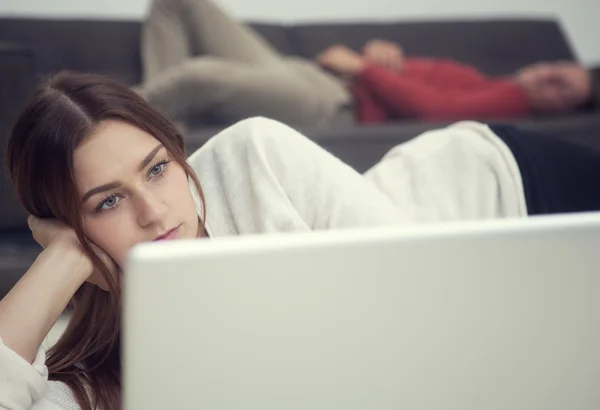 Młoda kobieta, patrząc na laptopa i człowieka leżącego na kanapie w backgrou — Zdjęcie stockowe