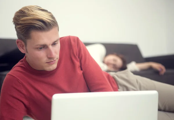 Blond man looking at laptop and woman on couch in background — Stock Photo, Image