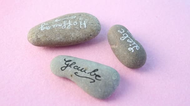 Três pedras com palavras alemãs para Amor, Acredite e Esperança em fundo rosa rotativo — Vídeo de Stock