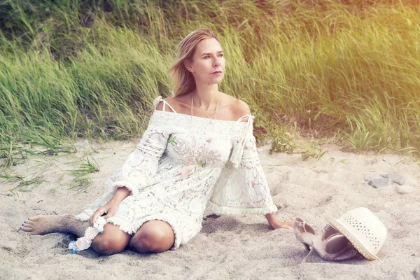 Mujer rubia en vestido blanco sentada en la playa — Foto de Stock