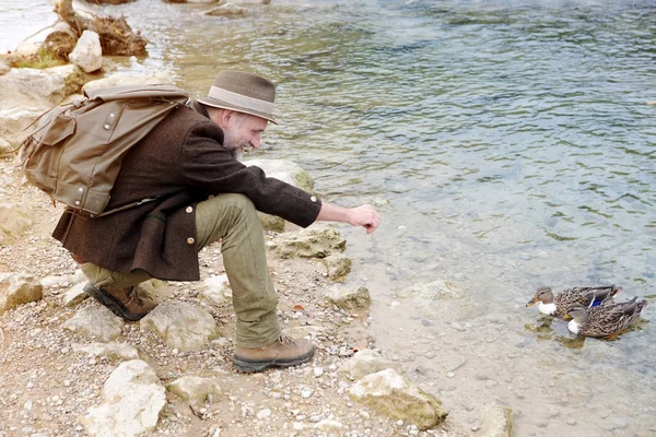 Homem em seus 50 anos sentado junto à água e alimentando patos — Fotografia de Stock