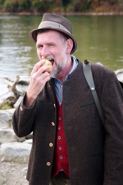 Bavarian man standing by river and eating apple — Stock Photo, Image