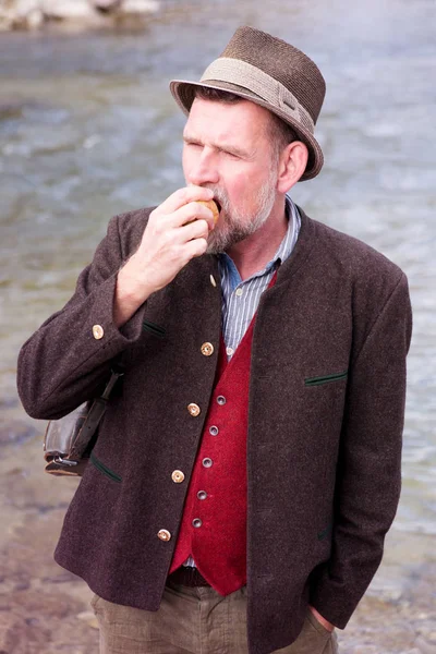 Bavarian man standing by river and eating apple — Stock Photo, Image