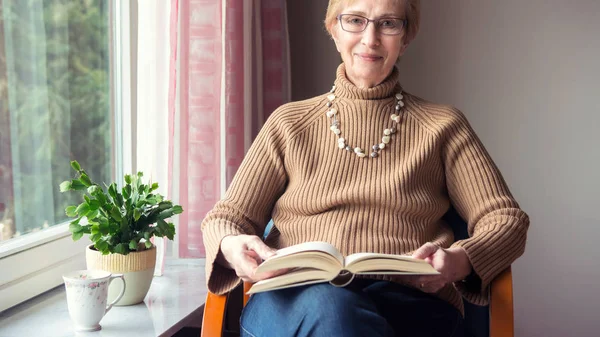 Cam kenarında oturan ve bir boo okuma yaşlı kadın — Stok fotoğraf