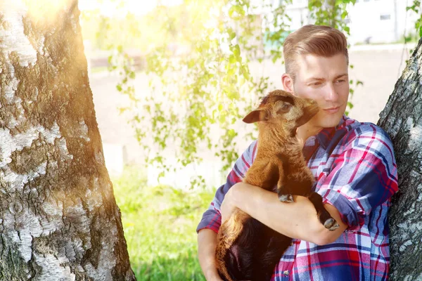 Blond man holding a baby goat next to a tree — Stock Photo, Image