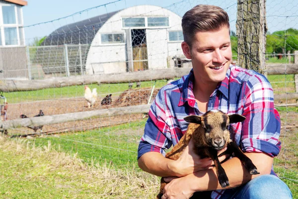 Blond man holding a baby goat in front of a barn — Stock Photo, Image