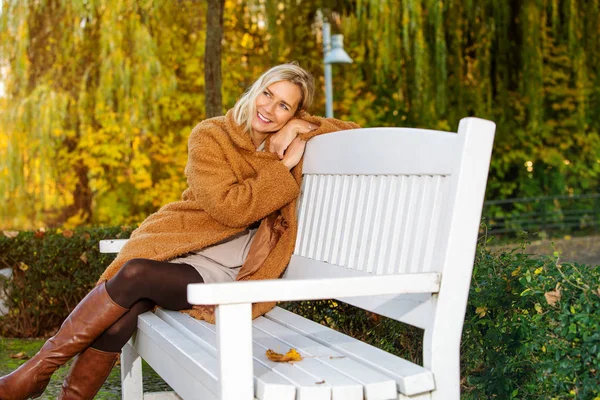 Femme blonde assise sur un banc dans le parc et profitant du soleil d'automne — Photo