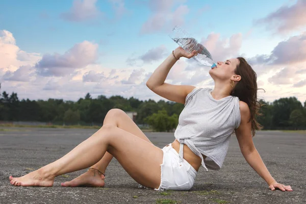 年轻女人穿着短裤坐在路面和饮用水 — 图库照片