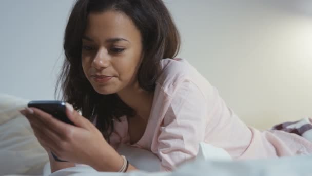 Porträt einer lächelnden jungen Afroamerikanerin, die mit Handy im Bett liegt. — Stockvideo