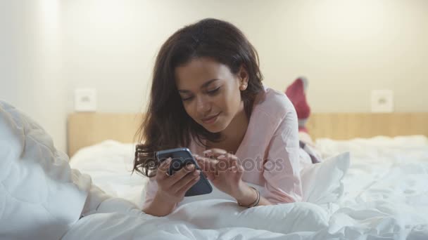 Portret van een lachende jonge Afro-Amerikaanse vrouw liggend in bed met mobiele telefoon. — Stockvideo