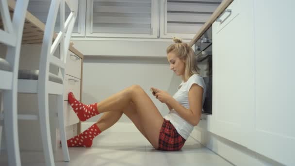 Giovane donna bionda seduta in un pavimento della cucina e digitando sul telefono cellulare . — Video Stock