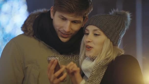 Νεαρό ζευγάρι βλέποντας φωτογραφίες στο smartphone το βράδυ σε ένα δρόμο της πόλης. — Αρχείο Βίντεο